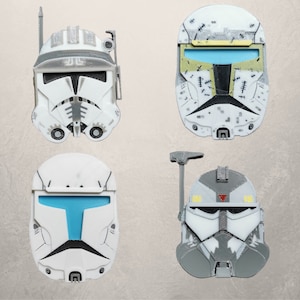 May include: Four white plastic Star Wars clone trooper helmets. The helmets have different designs and details. One helmet has a blue visor, another has a yellow visor, and the other two have clear visors. The helmets are all against a white background.