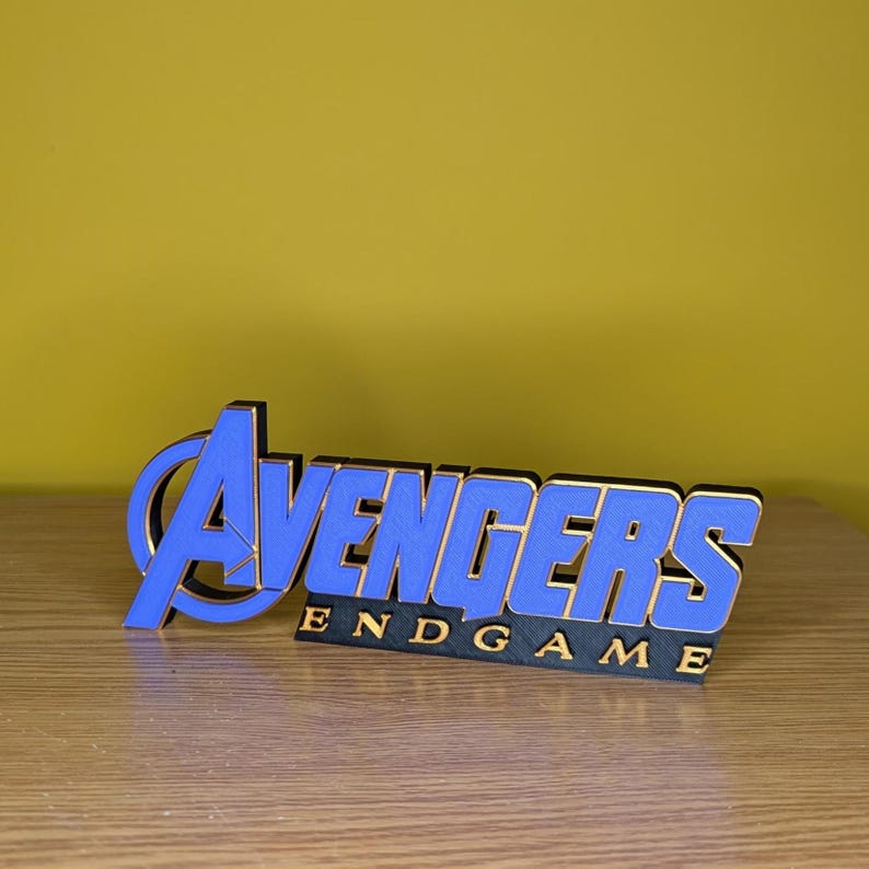 May include: A blue and gold Avengers Endgame logo, 3D printed and displayed on a wooden surface. The word "Avengers" is in blue with a gold outline, and "Endgame" is in gold lettering on a black base.