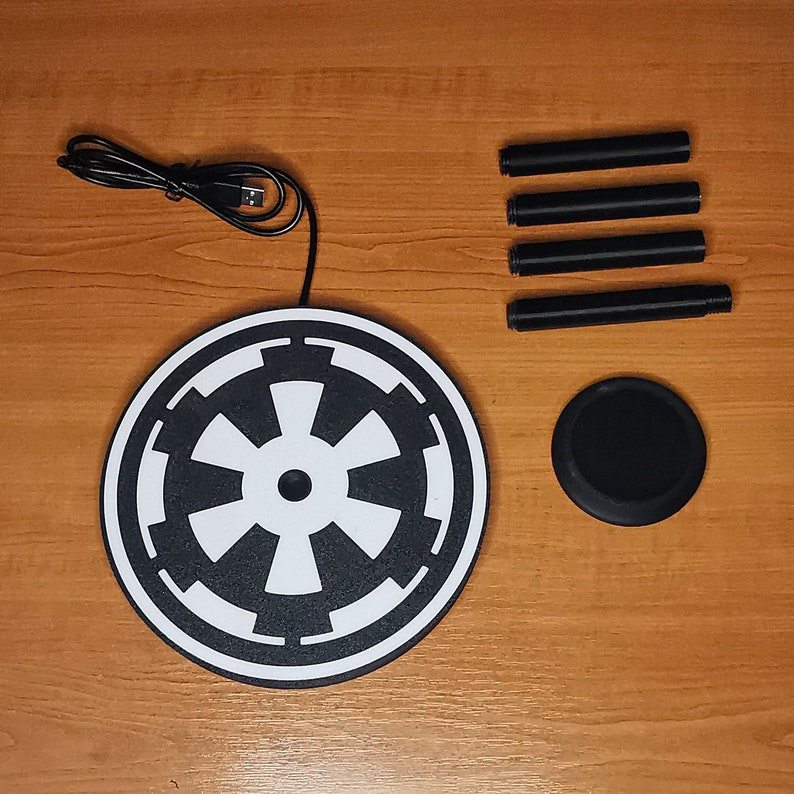 May include: A black and white wireless charging pad with a Star Wars Imperial symbol design. The pad is on a wooden surface with four black charging cables.