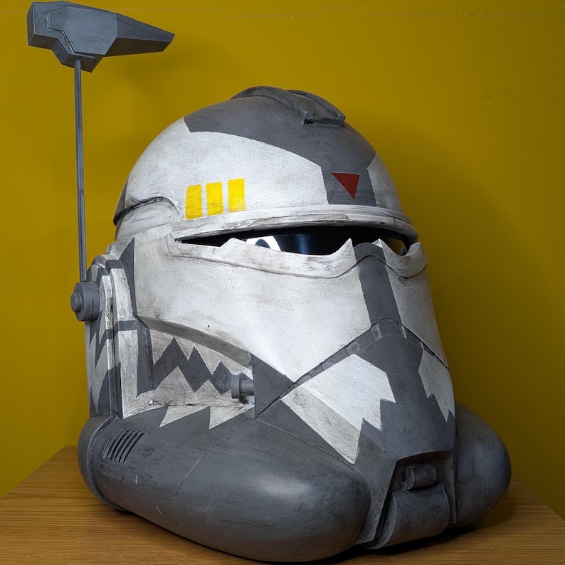 May include: A white and gray helmet with a black and white camouflage pattern. The helmet has a yellow stripe and a red triangle on the top. It has a black antenna on top.