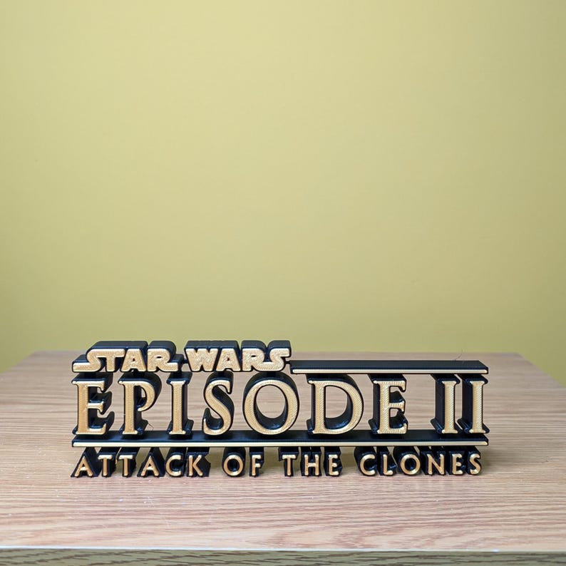 May include: A black and gold 3D printed sign that reads "STAR WARS EPISODE II ATTACK OF THE CLONES".
