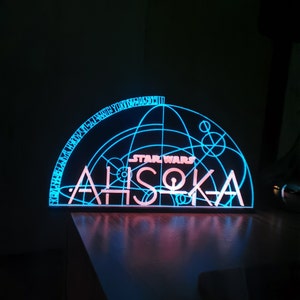 May include: A blue and pink LED light with a Star Wars Ahsoka Tano design. The light features a geometric pattern and the text "STAR WARS" and "AHSOKA".