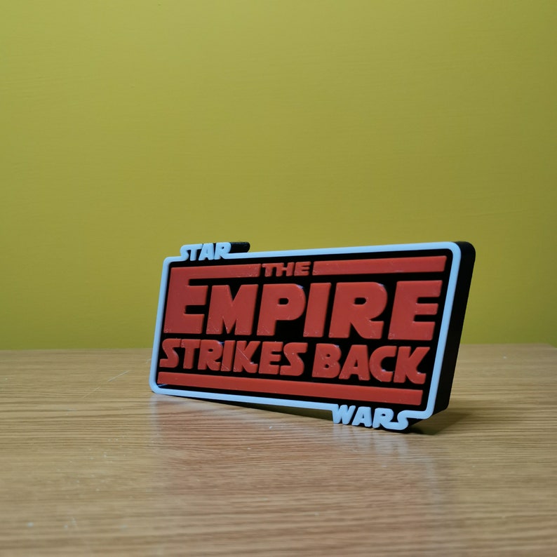 May include: A black and white 3D printed sign with the text "STAR WARS THE EMPIRE STRIKES BACK" in red and white letters.