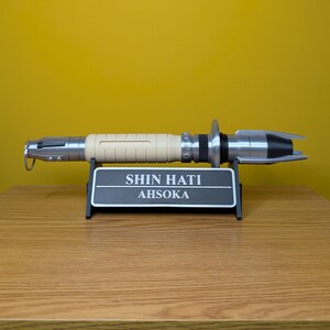 May include: A white and silver lightsaber with a black stand. The stand has a black plaque with the text "SHIN HATI AHSOKA" on it.