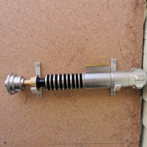 May include: A silver lightsaber with a black handle and a gold accent is mounted on a wall with a white bracket. The lightsaber is on a brown wood panel background.