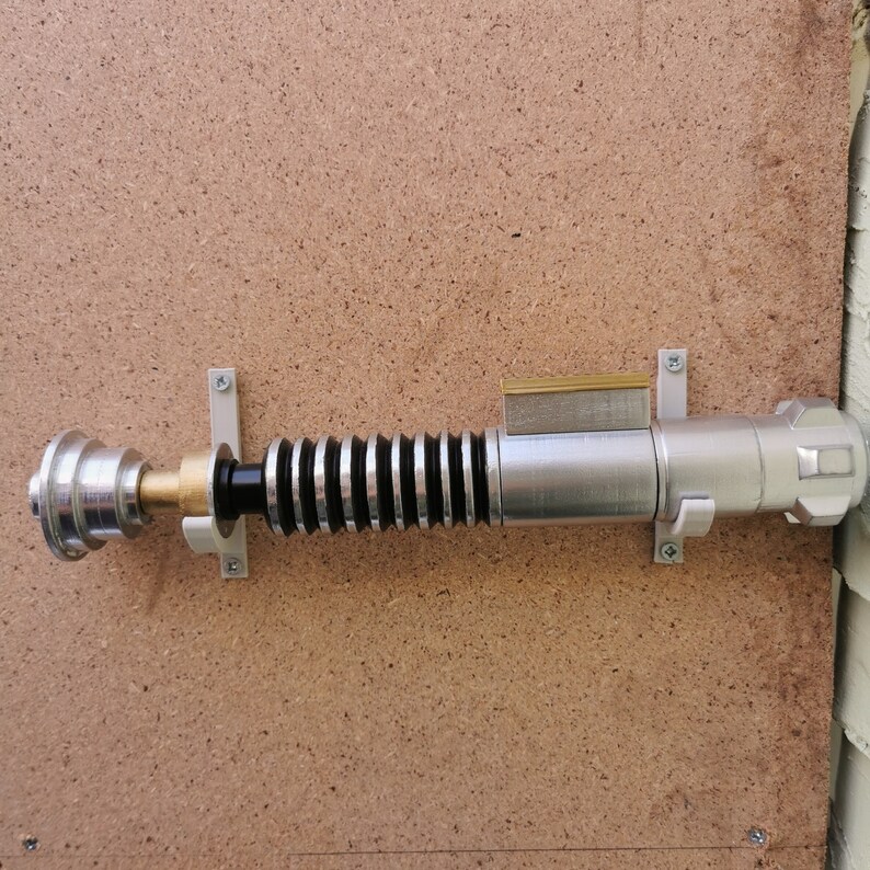 May include: A silver lightsaber with a black handle and a gold accent is mounted on a wall with a white bracket. The lightsaber is on a brown wood panel background.