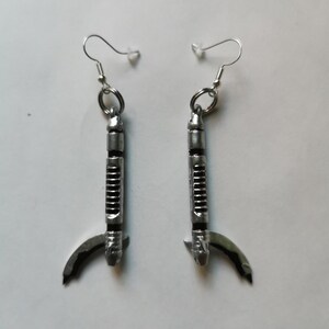 May include: A pair of silver earrings shaped like a lightsaber with a curved handle. The earrings are hanging from silver hooks.