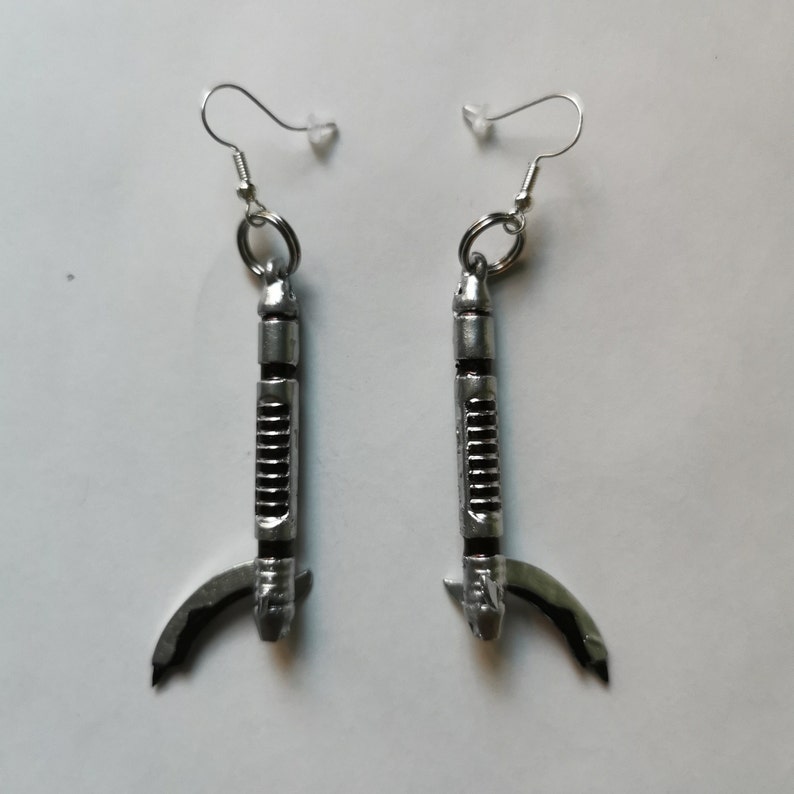 May include: A pair of silver earrings shaped like a lightsaber with a curved handle. The earrings are hanging from silver hooks.
