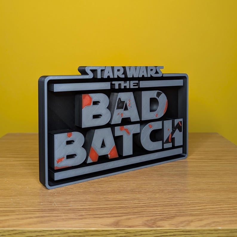 May include: A black and gray 3D printed sign with the text "STAR WARS THE BAD BATCH" in white letters with red accents.