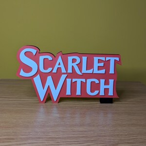 May include: A red and white sign with the words "SCARLET WITCH" in a bold, layered design. The sign is set against a light brown wooden surface and a yellow background.