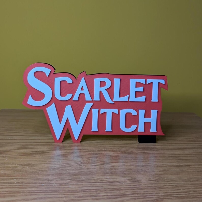 May include: A red and white sign with the words "SCARLET WITCH" in a bold, layered design. The sign is set against a light brown wooden surface and a yellow background.
