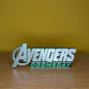 以下が含まれることがあります： 銀と緑で「Avengers」、緑と黒で「DOOMSDAY」と書かれた3Dプリントのサイン。 黄色の背景に、木製の表面に置かれています。