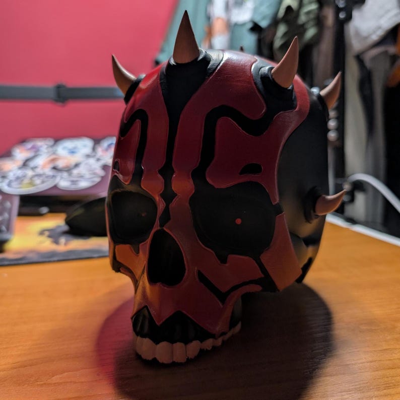 May include: A detailed, hand-painted skull mask in black and red, featuring prominent horns. The mask has a skull-like design with intricate patterns and is likely a costume or collectible item. The mask is displayed on a wooden surface.