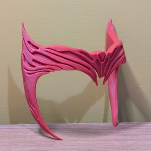May include: A red plastic headband with a detailed, textured design. The headband is curved and has two pointed ends.