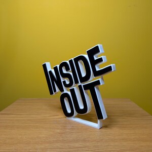 May include: White 3D printed sign with the words "Inside Out" in black letters. The sign is tilted to the side and is sitting on a wooden surface.