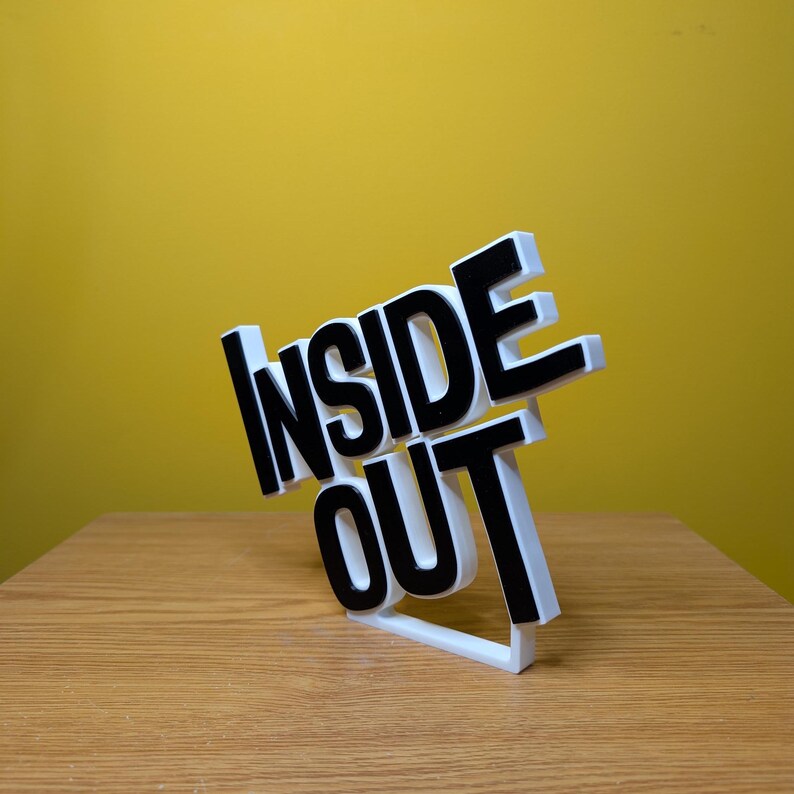 May include: White 3D printed sign with the words "Inside Out" in black letters. The sign is tilted to the side and is sitting on a wooden surface.