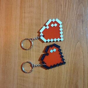 May include: Two red pixel heart keychains with black and white outlines. One keychain has a white outline and the other has a black outline.
