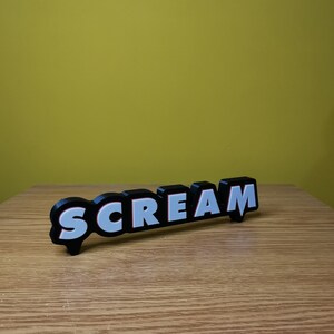 May include: Black and white 3D word sign that reads "SCREAM" with red outlines. The sign is on a wooden surface.