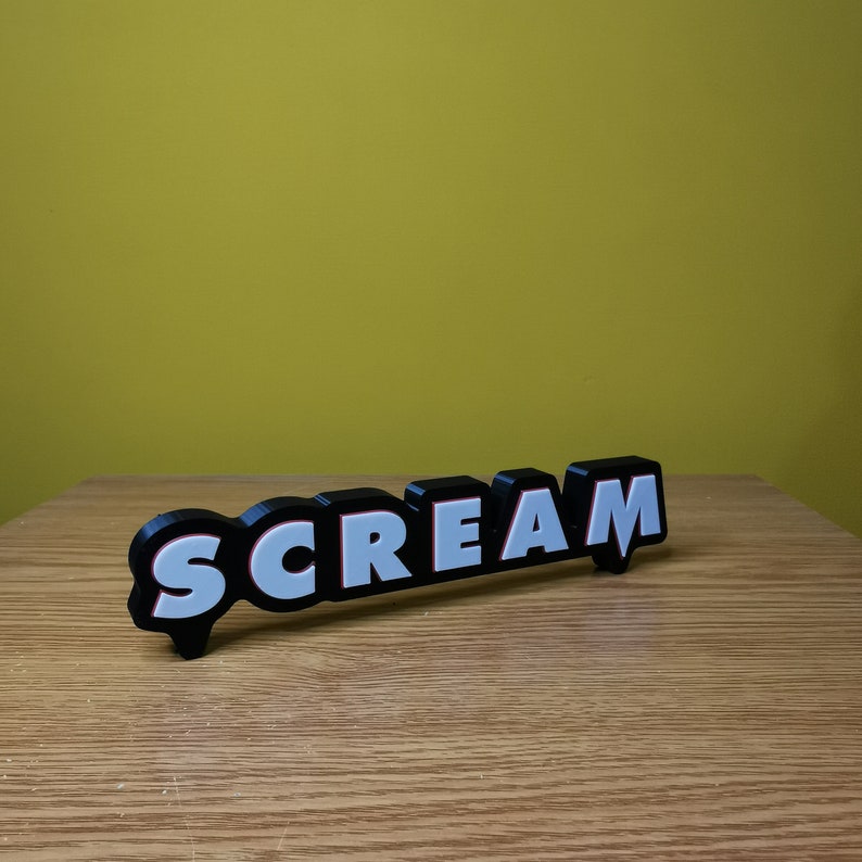 May include: Black and white 3D word sign that reads "SCREAM" with red outlines. The sign is on a wooden surface.