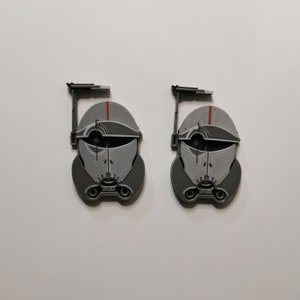 May include: Two gray and white 3D printed Star Wars clone trooper helmet figures with red accents. The helmets are facing forward and have a black antenna on top.
