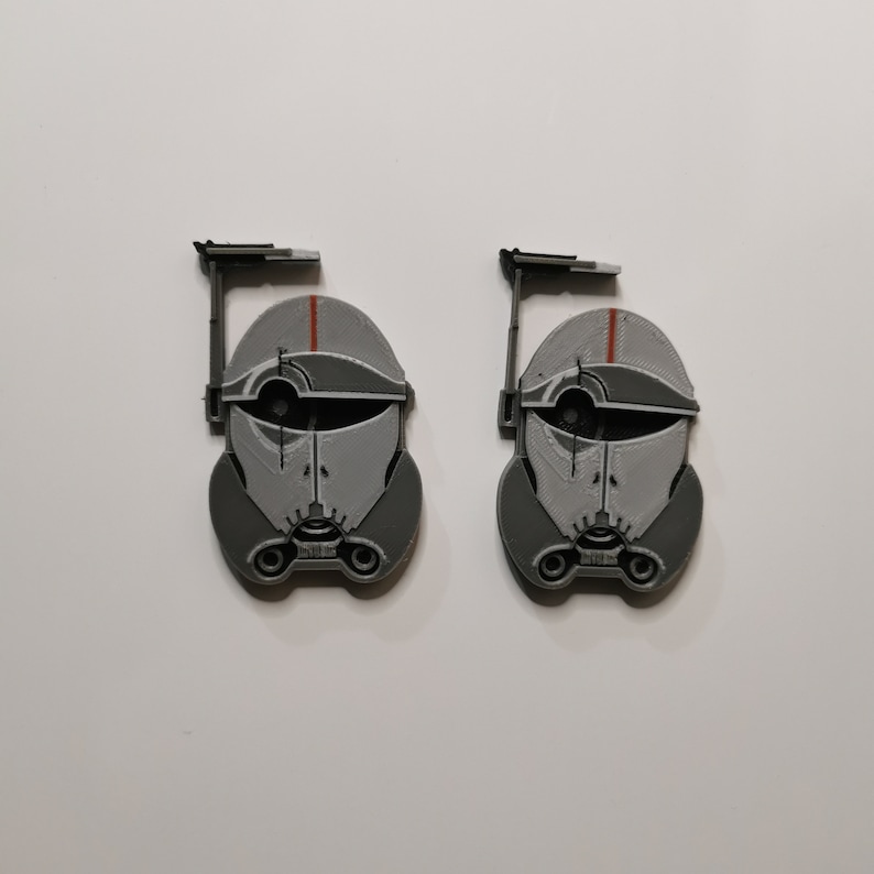 May include: Two gray and white 3D printed Star Wars clone trooper helmet figures with red accents. The helmets are facing forward and have a black antenna on top.