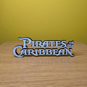 May include: White 3D printed sign with the text "Pirates of the Caribbean" in a black outline.
