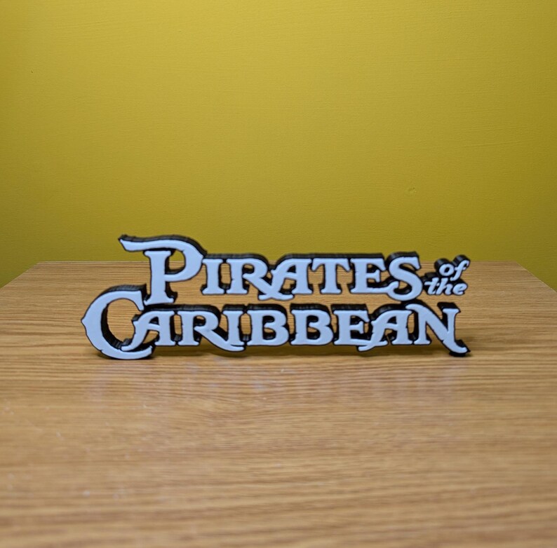 May include: White 3D printed sign with the text "Pirates of the Caribbean" in a black outline.