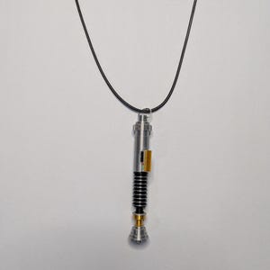 May include: A silver-colored lightsaber pendant with black and gold accents hangs from a black cord. The pendant has a detailed design with a cylindrical body, black ribbed section, and a gold-colored base. The pendant is suspended against a white background.