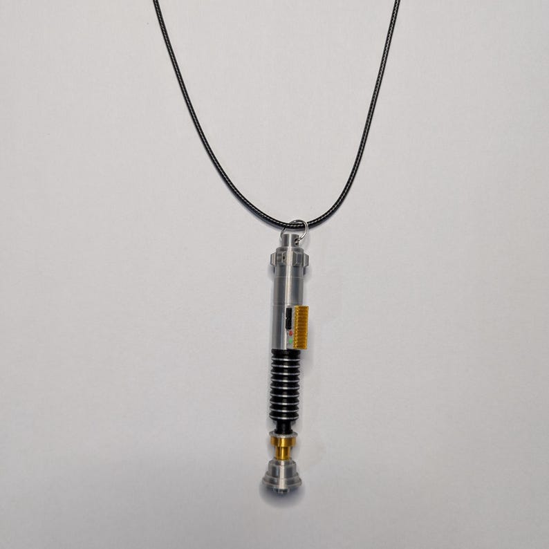 May include: A silver-colored lightsaber pendant with black and gold accents hangs from a black cord. The pendant has a detailed design with a cylindrical body, black ribbed section, and a gold-colored base. The pendant is suspended against a white background.