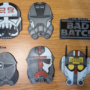May include: Six Star Wars: The Bad Batch character helmet designs. The helmets are white, black, gray, and red with various details. The helmets are made of plastic and are approximately 3 inches in diameter. The helmets are displayed on a wooden surface. The text "Star Wars The Bad Batch" is displayed on a black rectangle with a gray background.