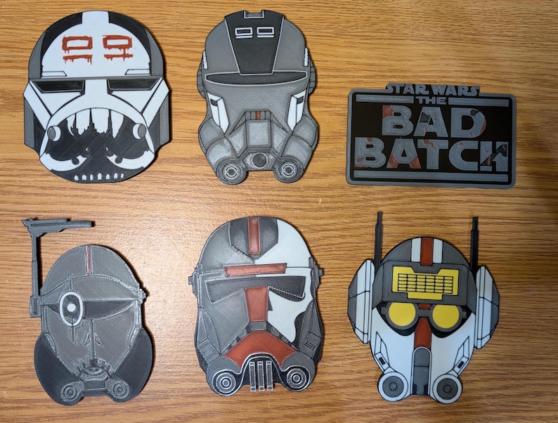 May include: Six Star Wars: The Bad Batch character helmet designs. The helmets are white, black, gray, and red with various details. The helmets are made of plastic and are approximately 3 inches in diameter. The helmets are displayed on a wooden surface. The text "Star Wars The Bad Batch" is displayed on a black rectangle with a gray background.