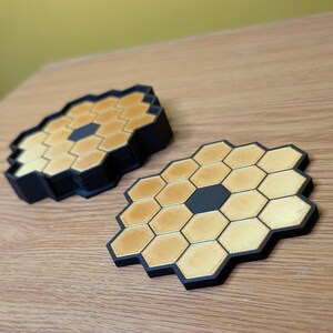 May include: Two black and gold honeycomb-shaped coasters. The coasters are made of a 3D printed material and have a hexagonal pattern.