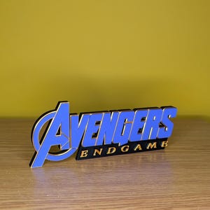 May include: A blue and gold Avengers Endgame logo, with the word "Avengers" in blue and the word "Endgame" in gold. The logo is set against a yellow background and sits on a wooden surface.