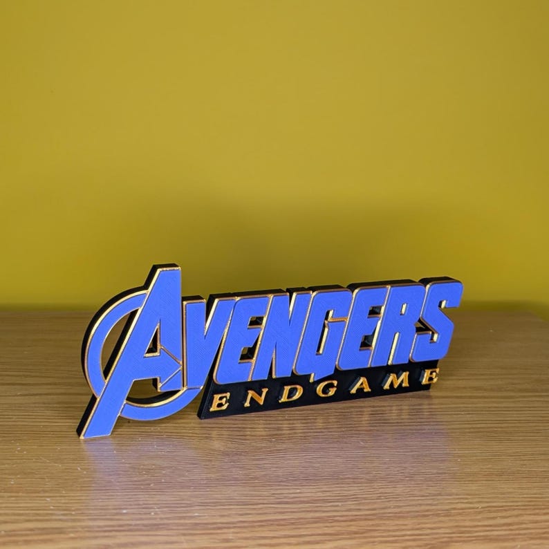 May include: A blue and gold Avengers Endgame logo, with the word "Avengers" in blue and the word "Endgame" in gold. The logo is set against a yellow background and sits on a wooden surface.