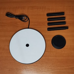 May include: A white wireless charging pad with a black charging coil in the center. The pad is surrounded by a black ring. The charging pad is on a wooden surface. There are four black charging cables on the surface.