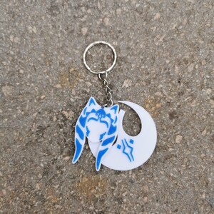 Pu&ograve; includere: Un portachiavi bianco e blu con un'immagine stilizzata di Ahsoka Tano, un personaggio di Star Wars, con una forma di mezzaluna dietro di lei.