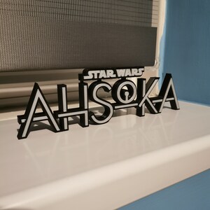 May include: A black and white 3D-printed sign that reads "STAR WARS AHSOKA". The sign is placed on a white surface, with a window shade in the background. The letters are black with white outlines.