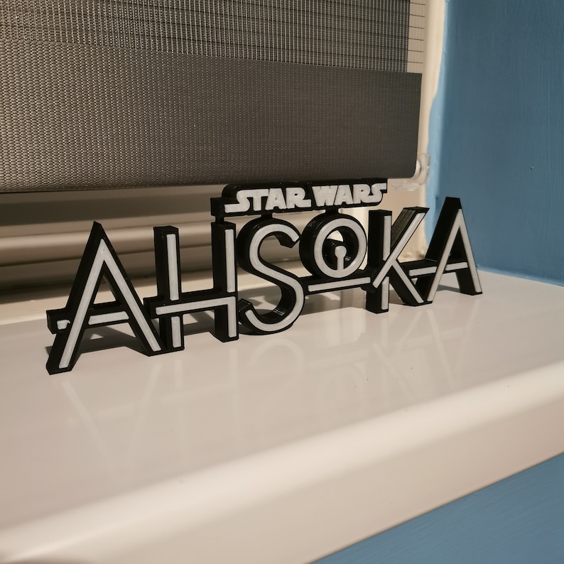 May include: A black and white 3D-printed sign that reads "STAR WARS AHSOKA". The sign is placed on a white surface, with a window shade in the background. The letters are black with white outlines.