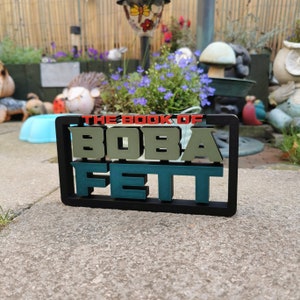 Peut inclure: Un panneau noir encadr&eacute; avec le texte "The Book of Boba Fett" en lettres vertes et rouges. Le panneau est pos&eacute; sur une surface en b&eacute;ton gris.