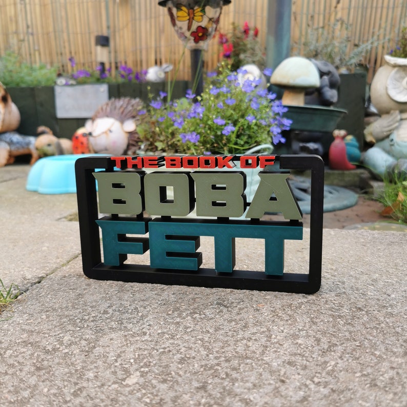 Peut inclure: Un panneau noir encadr&eacute; avec le texte "The Book of Boba Fett" en lettres vertes et rouges. Le panneau est pos&eacute; sur une surface en b&eacute;ton gris.