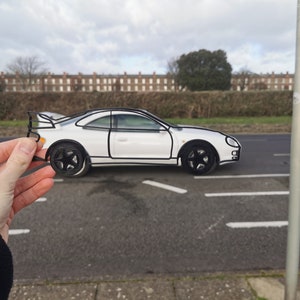 May include: A white car drawn in black outline is held in a person's hand. The car is a coupe with a spoiler and large wheels. The car is in front of a blurry background of a street and buildings.