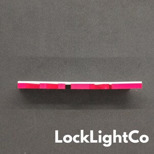 Op de afbeelding: Een wit en rood rechthoekig object met een reeks rode blokken langs de onderrand. Het object staat tegen een donkergrijze achtergrond. De tekst "LockLightCo" staat onderaan.