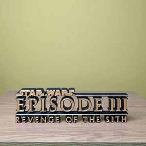 May include: A black and gold 3D printed sign that reads "Star Wars Episode III Revenge of the Sith".