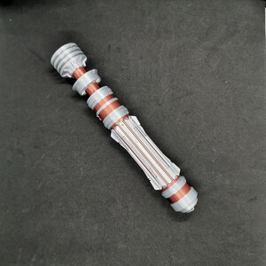 May include: A silver and copper lightsaber prop with a detailed design. The lightsaber is made of plastic and has a textured surface.
