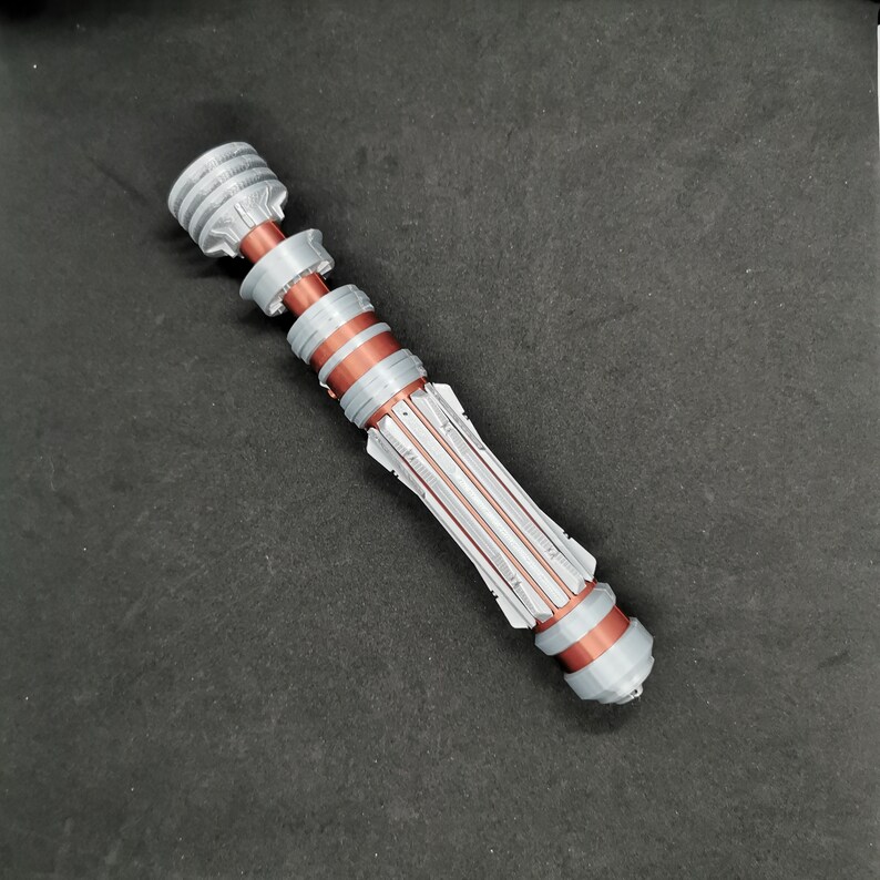 May include: A silver and copper lightsaber prop with a detailed design. The lightsaber is made of plastic and has a textured surface.