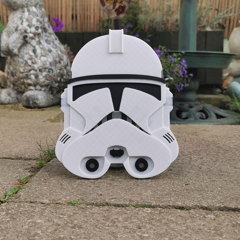 May include: A white and black Stormtrooper helmet replica, featuring the iconic design from the Star Wars franchise. The helmet has black eye and mouth details, with ventilation slits on the sides. The helmet is made of a hard material.