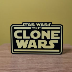 以下が含まれることがあります： 「スター・ウォーズ クローン・ウォーズ」の文字が書かれた黒と黄色の3Dプリントされた看板です。