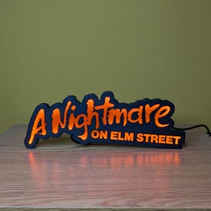May include: A black and orange illuminated sign that reads "A Nightmare on Elm Street." The sign has a black border with orange lettering and a glowing orange interior. The sign is on a wooden surface.