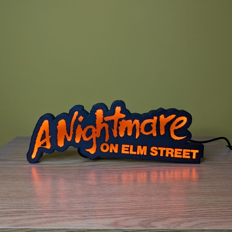 May include: A black and orange illuminated sign that reads "A Nightmare on Elm Street." The sign has a black border with orange lettering and a glowing orange interior. The sign is on a wooden surface.