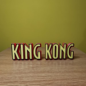 Pu&ograve; includere: Lettere di plastica rosse e nere che compongono la scritta "KING KONG" su una superficie di legno.